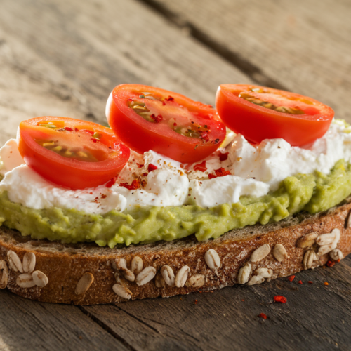 A vibrant and appetizing slice of whole grain toast topped with creamy cottage cheese and mashed avocado, garnished with sliced cherry tomatoes and red pepper flakes, on a rustic wooden table with a bright morning light.