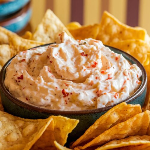 A close-up of a bowl of creamy, spicy cottage cheese dip with flecks of red chili, surrounded by golden, crispy tortilla chips. Background features a casual kitchen setting with warm tones.